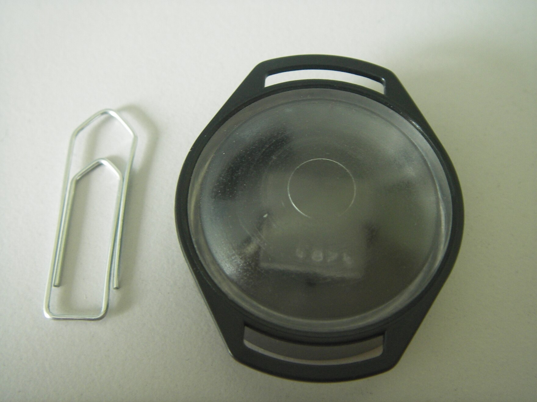 Die Form des Dosimeters ähnelt einer Armbanduhr. Die Größe wird durch eine etwa gleich große Büroklammer verdeutlicht.