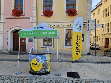 Stand der mobilen Radonberatung. Unter einem grün-weißen Pavilion steht ein Beratungstresen und neben dem Pavillion eine Beachflag mit der Aufschrift "Radonberatung"