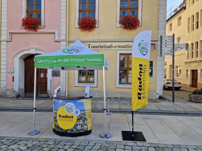 Stand der mobilen Radonberatung. Unter einem grün-weißen Pavilion steht ein Beratungstresen und neben dem Pavillion eine Beachflag mit der Aufschrift "Radonberatung"
